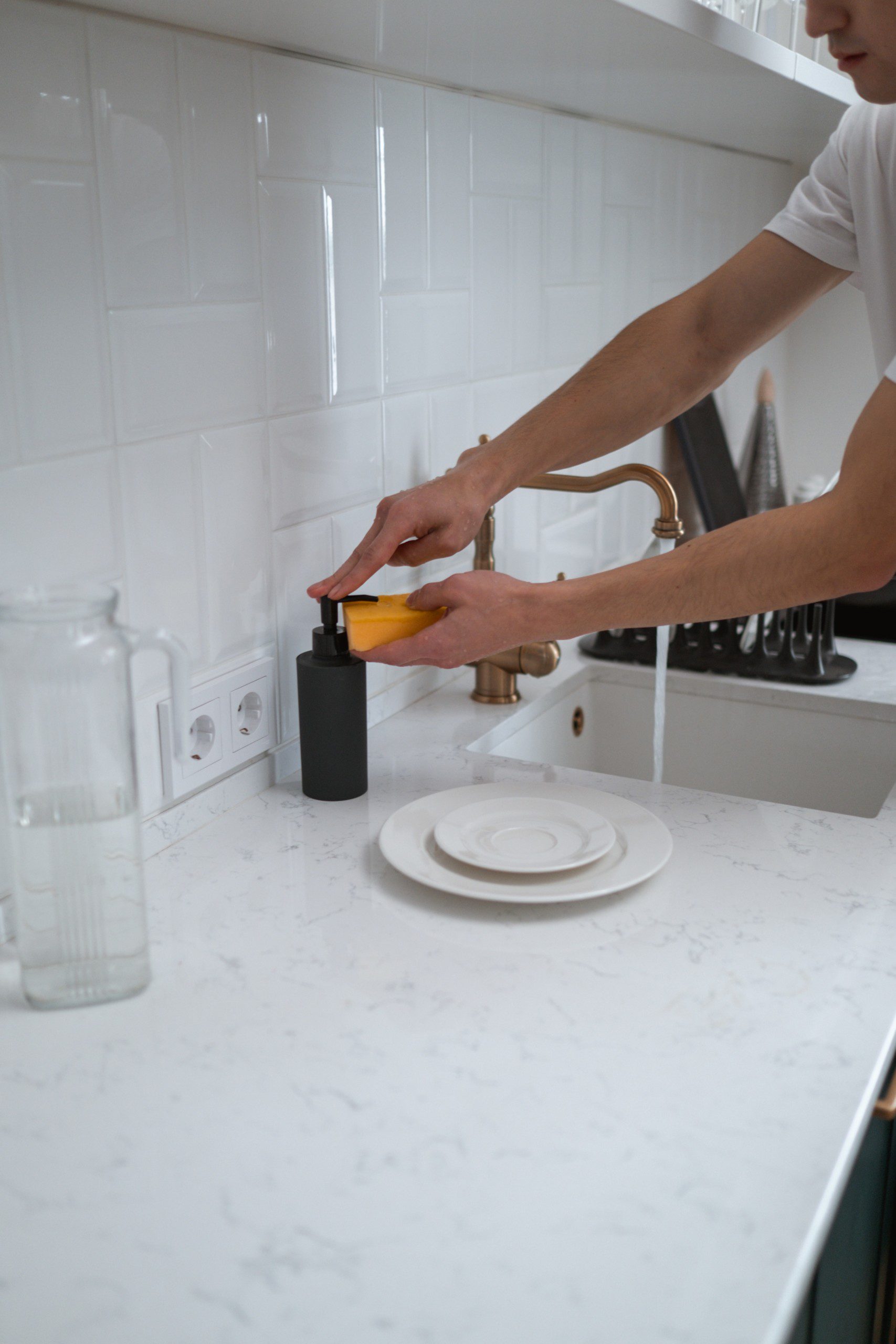 Imagem de uma cozinha com azulejos brancos e uma pessoa lavando um prato de porcelana branca com detergente caseiro.