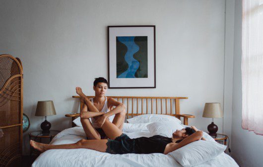 A imagem contém duas mulheres deitadas em uma cama no quarto de casal. Uma está de camiseta regata branca e a outra preta. No quarto há um quadro na parede ao fundo da cama, uma cabeceira de madeira, uma mesinha de cabeceira de cada lado da cama com abajur.