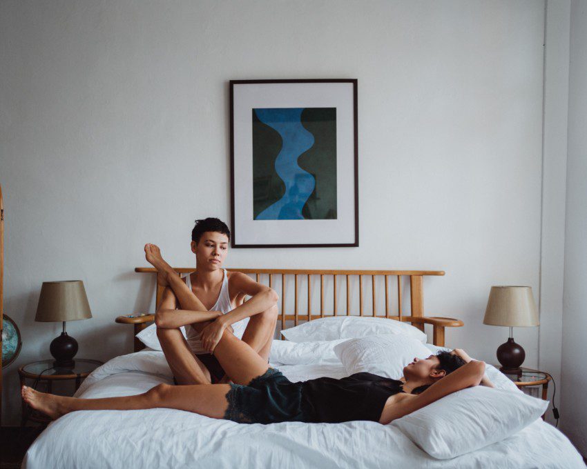 A imagem contém duas mulheres deitadas em uma cama no quarto de casal. Uma está de camiseta regata branca e a outra preta. No quarto há um quadro na parede ao fundo da cama, uma cabeceira de madeira, uma mesinha de cabeceira de cada lado da cama com abajur.