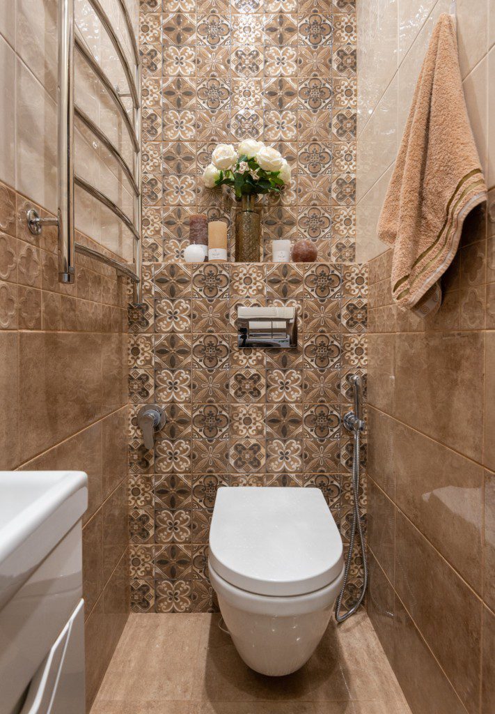 Imagem de um lavabo decorado com azulejos estilizados no tom de marrom, um vaso sanitário branco e moderno no fundo, uma prateleira com um vasinho de flor e algumas velas e no canto, uma pia branca.