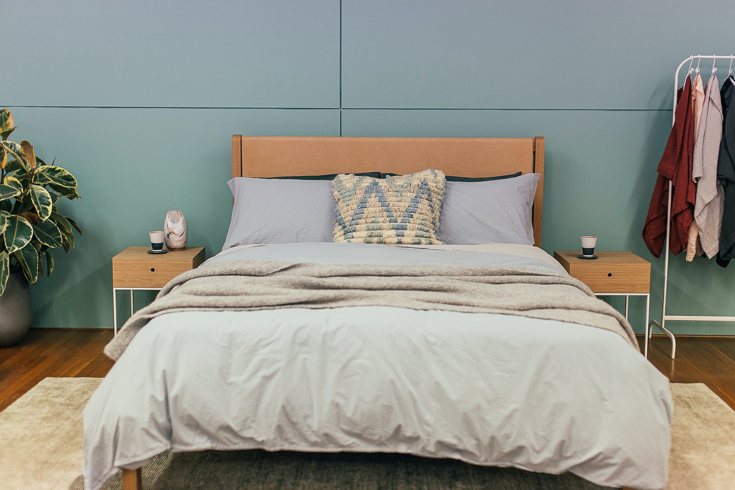 Imagem de um quarto de casal com cama e cabeceira de madeira. As mesinhas de cabeceira também são de madeira. O quarto possui uma parede no fundo no tom verde escuro e uma planta no canto esquerdo da foto.