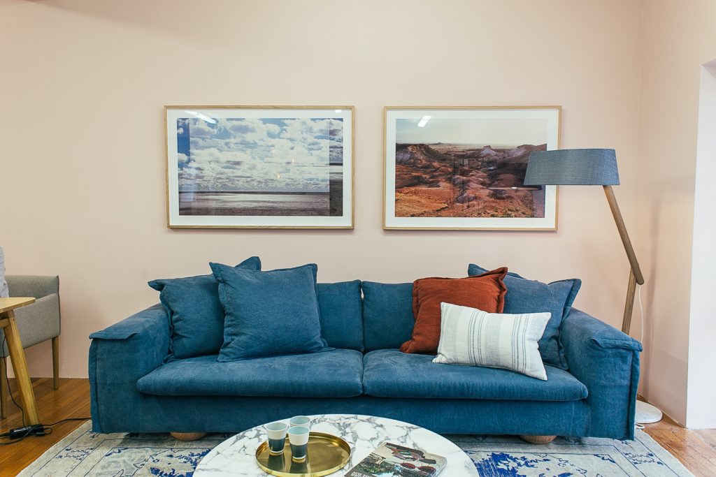 Imagem de uma sala decorada com 2 quadros na parede, um sofá azul, uma luminária de chão no canto direito e no centro, um tapete branco com azul e uma mesa de centro redonda de mármore.