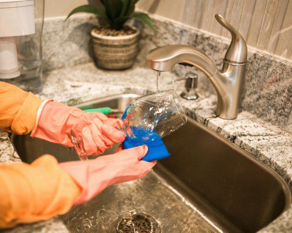 Imagem de uma pessoa lavando a louça. É possível ver uma pia de mármore com uma plantinha no canto e uma pessoa limpando uma taça de vidro.