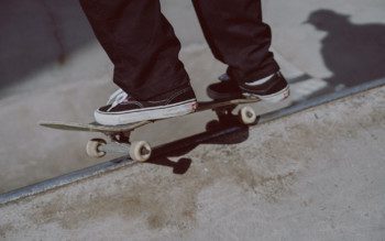 Foto que ilustra matéria sobre Pista de skate em Santos mostra um detalhe de um skate sendo equilibrado na borda de uma pista, com dois pés de um skatista sobre ele. O skatista usa calças compridas pretas e um tênis preto com parte do solado branco
