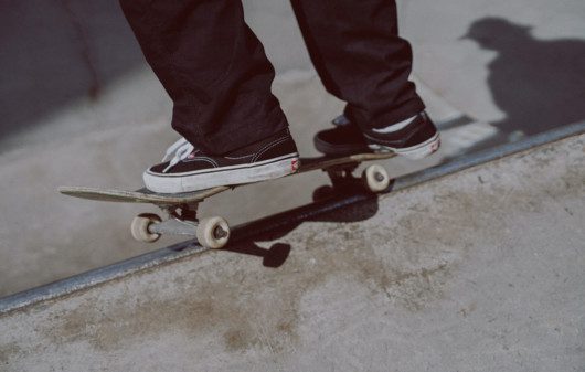 Foto que ilustra matéria sobre Pista de skate em Santos mostra um detalhe de um skate sendo equilibrado na borda de uma pista, com dois pés de um skatista sobre ele. O skatista usa calças compridas pretas e um tênis preto com parte do solado branco