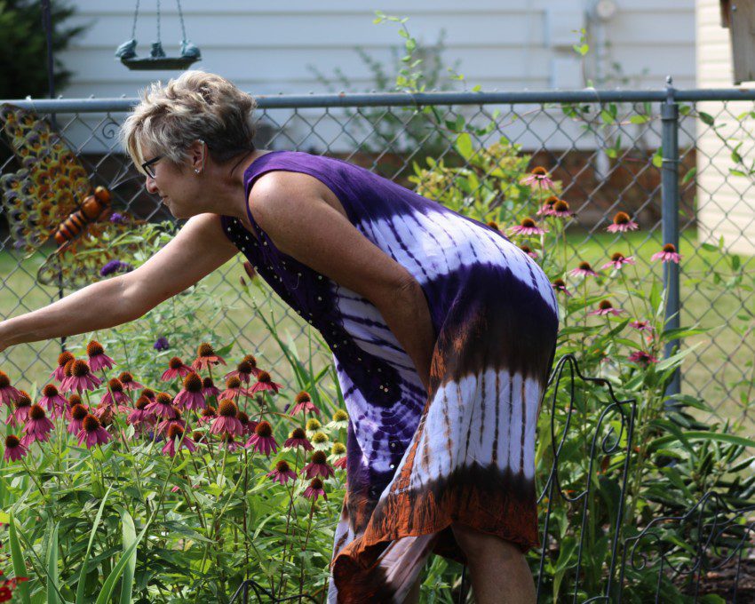 Foto que ilustra matéria sobre plantas para jardim mostra uma mulher de vestido roxo e branco estilo tie dye se aproximando de flores em seu jardim em casa.