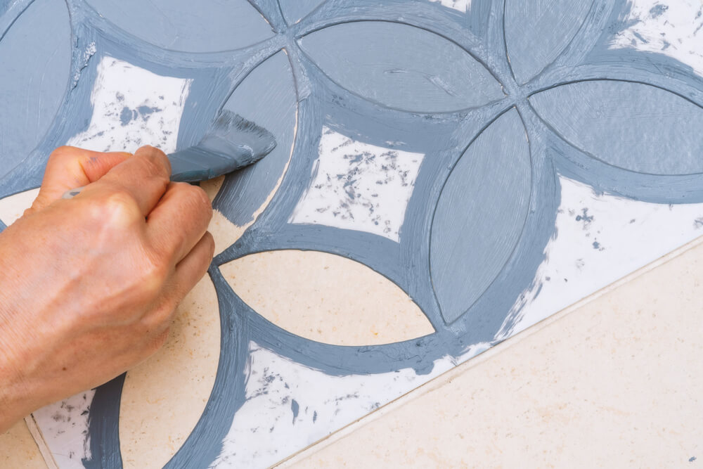 Foto que ilustra matéria sobre como fazer stencil mostra detalhes da pintura de um piso com um molde e um pincel grosso.