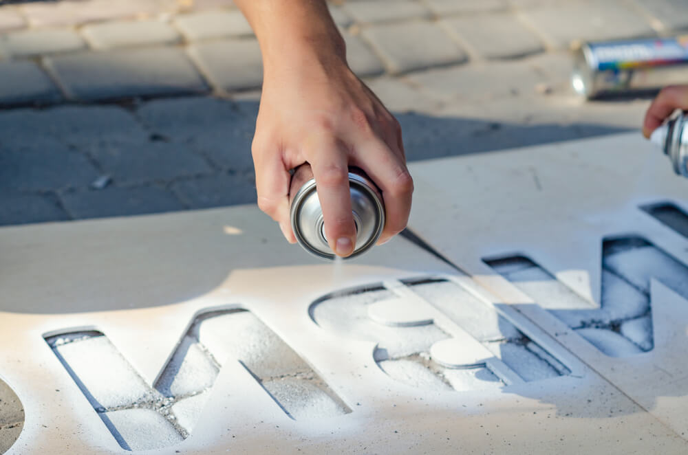 Foto que ilustra matéria sobre como fazer stencil mostra o uso de um molde para uma pintura de uma palavra no chão com o uso de tinta spray.
