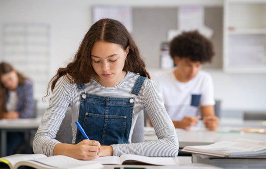 Foto que ilustra matéria sobre escolas em Santos mostra uma adolescentes de cabelos compridos pretos e cacheados, usando um macacão jeans com uma blusa cinza de mangas compridas por baixo, sentada a uma carteira de escola escrevendo em um caderno.