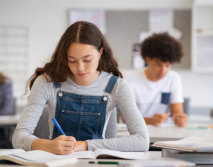 Foto que ilustra matéria sobre escolas em Santos mostra uma adolescentes de cabelos compridos pretos e cacheados, usando um macacão jeans com uma blusa cinza de mangas compridas por baixo, sentada a uma carteira de escola escrevendo em um caderno.