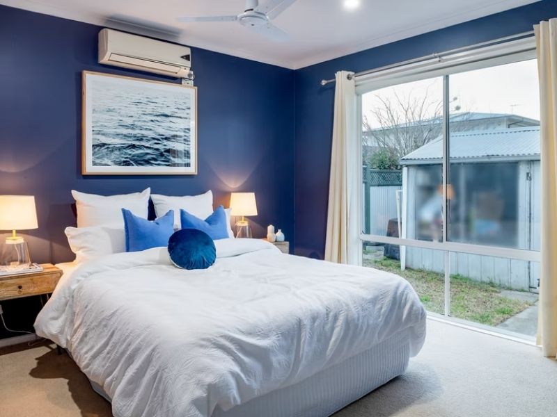 A foto mostra um quarto de casal com cores brancas e azuis. Há na imagem: uma cama de casal, duas mesas de cabeceira com abajur, um quadro, almofadas, travesseiros, ar condicionado, ventilador de teto e uma ampla janela lateral.