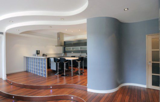 Foto que ilustra matéria sobre decoração de paredes curvas mostra a entrada de uma sala com chão de madeira e uma parede curva pintada de azul. Ao lado dela, uma cozinha americana, com uma mesa e banquetas.