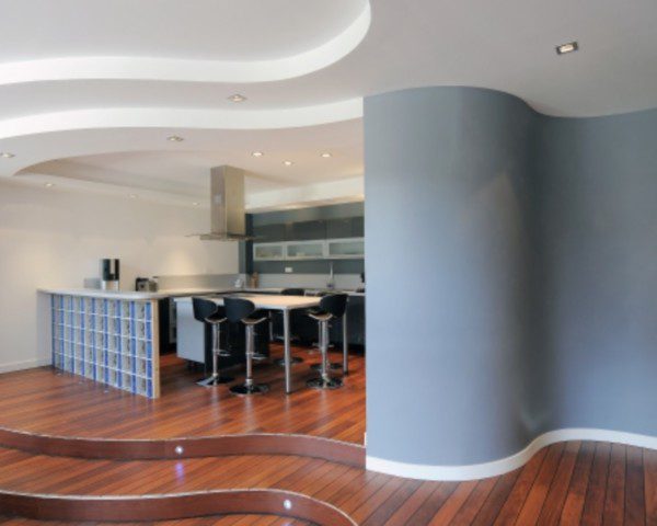 Foto que ilustra matéria sobre decoração de paredes curvas mostra a entrada de uma sala com chão de madeira e uma parede curva pintada de azul. Ao lado dela, uma cozinha americana, com uma mesa e banquetas.