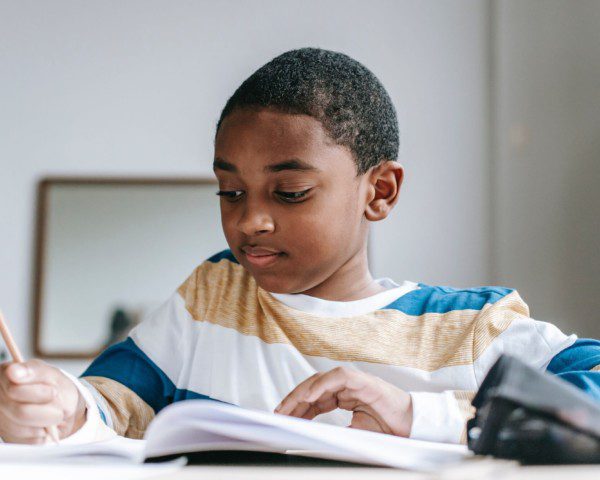 Foto que ilustra matéria sobre escolas em Santo André mostra um menino negro sentado à uma mesa enquanto escreve de lápis em um caderno.