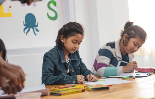 Foto que ilustra matéria sobre escolas em São Caetano do Sul mostra duas meninas, sentadas lado a lado à uma mesa enquanto fazem desenhos em folhas de papel.