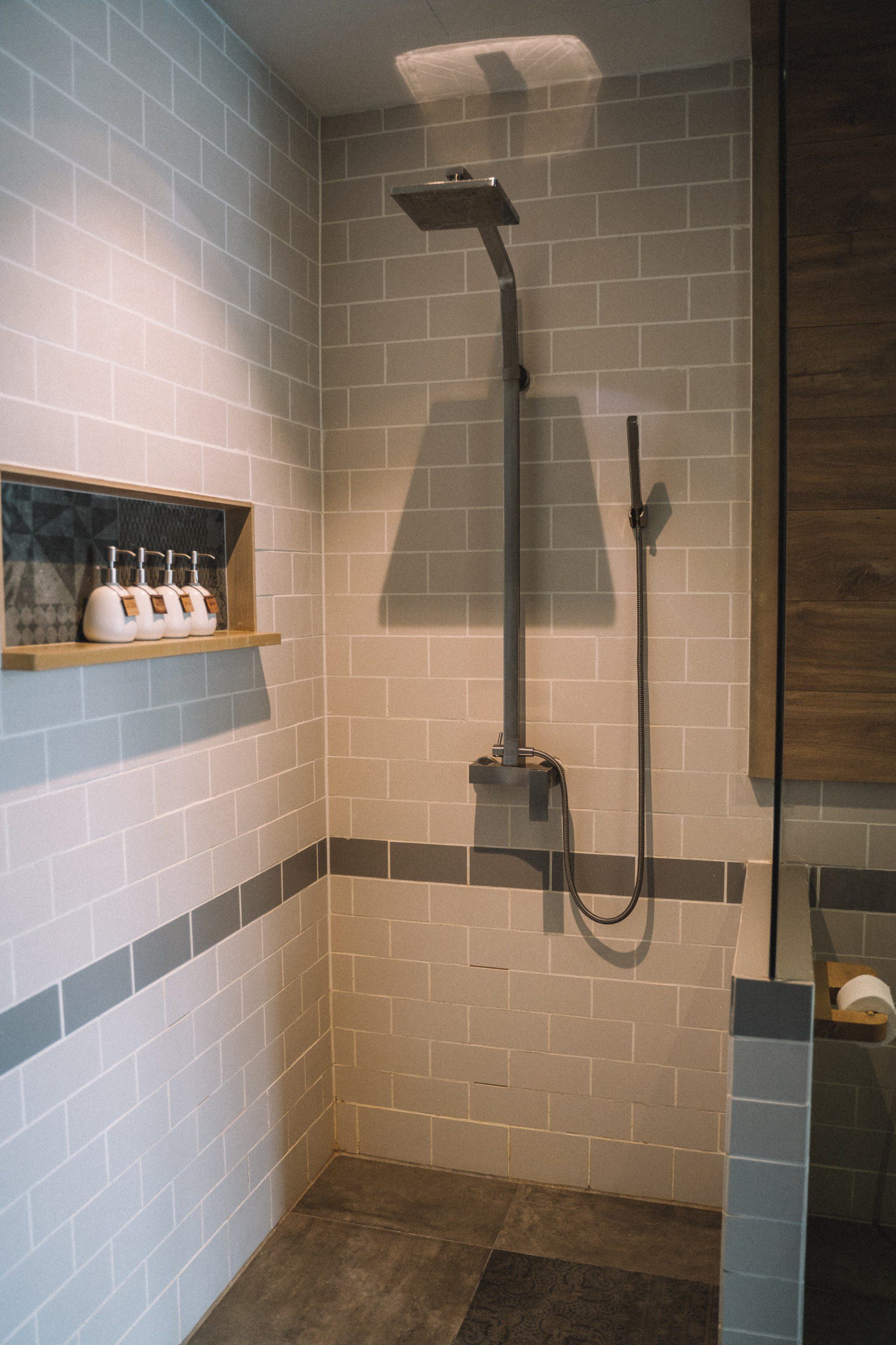 Imagem de um lavabo com um chuveiro de aço, uma parede com azulejos de tijolinhos e espaços com shampoos
