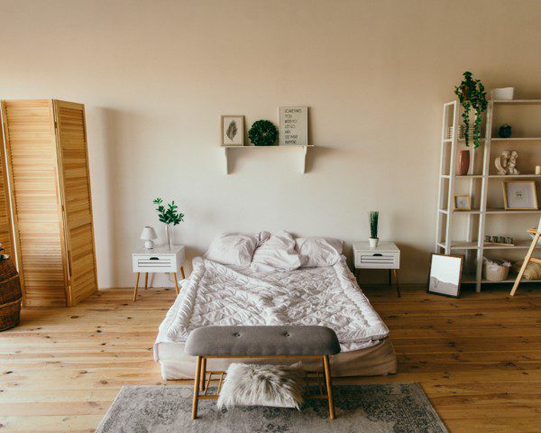 Imagem de um quarto grande com uma tela dobrável de madeira, uma cama de casal no centro e no canto direito uma estante com alguns objetos e plantas.
