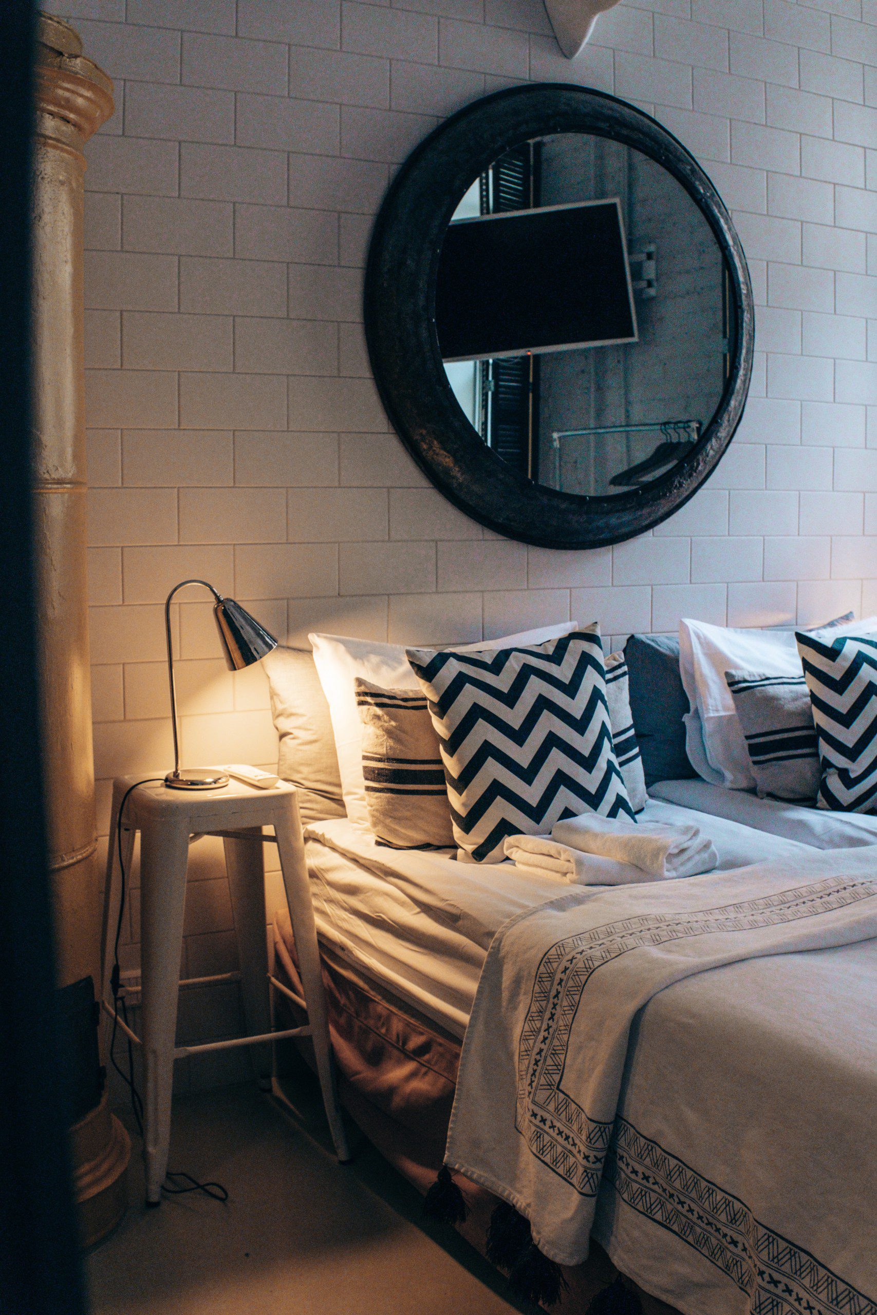 Cama de casal decorada com jogo de cama em preto e branco, almofadas estampadas na mesma cor, com espelho redondo atrás do móvel. 