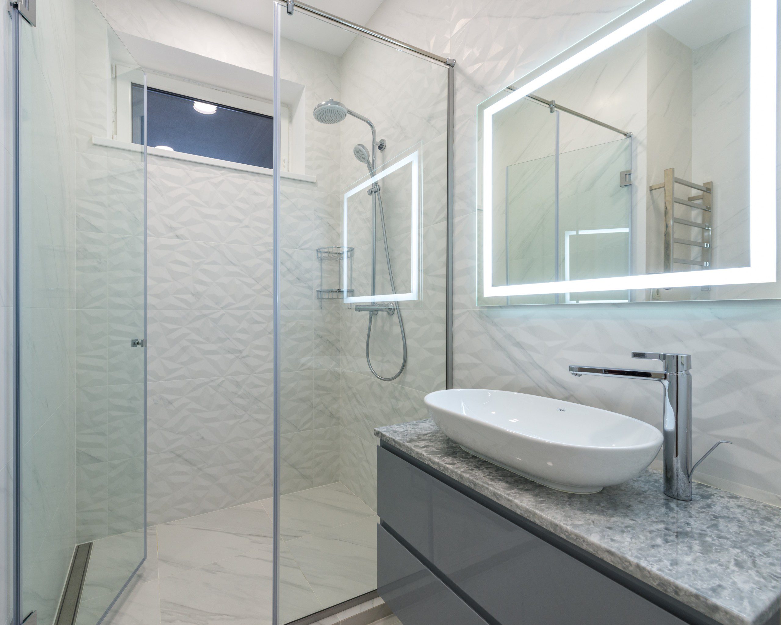 Imagem de um banheiro cinza com uma pia e o tampo de pedra de mármore, box de vidro e um chuveiro de aço inox