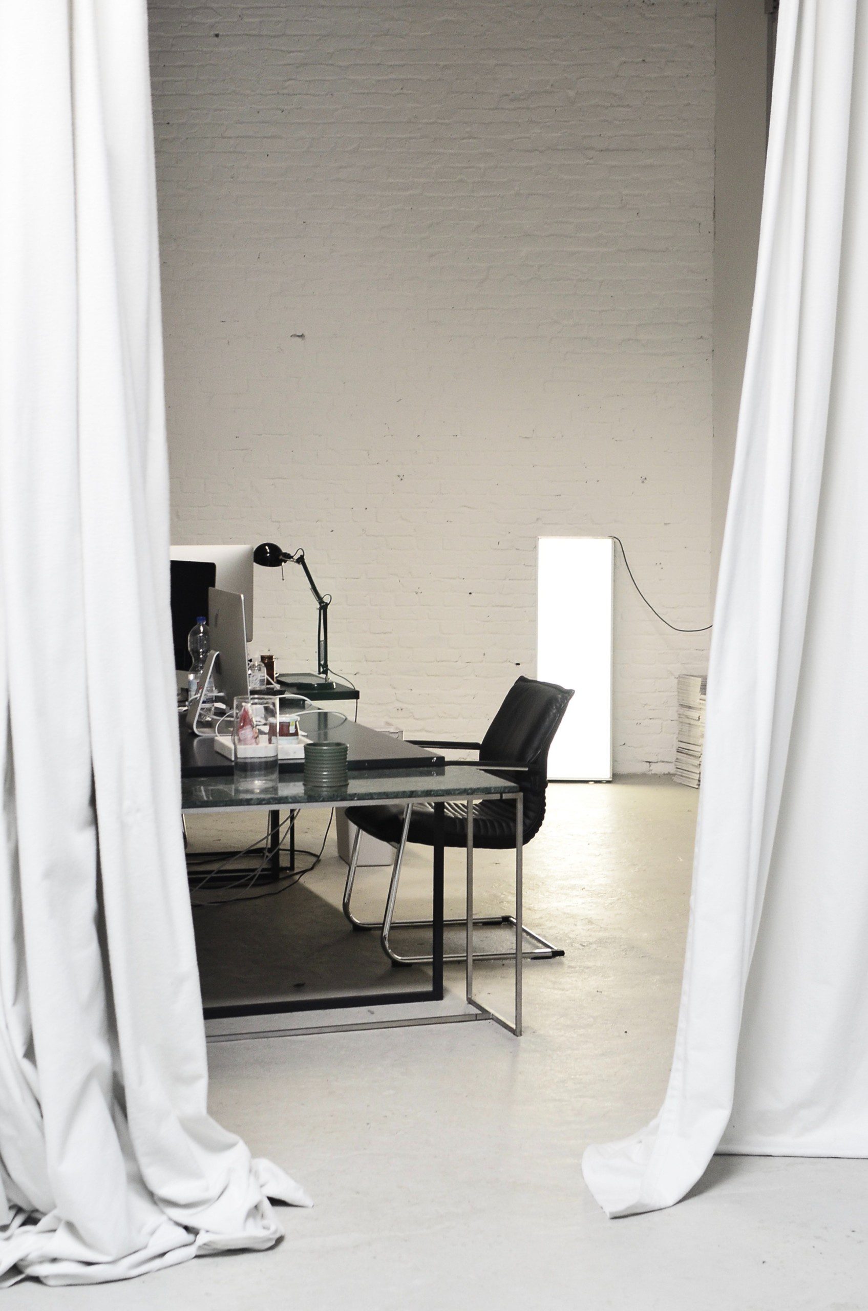 Imagem de um ambiente grande separado por um cortina branca. Ao fundo, uma mesa de escritório de aço com alguns objetos de estudo.