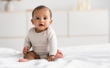Foto que ilustra matéria sobre quarto de bebê simples mostra um bebê de poucos meses sentado em cima de uma cama olhando para a câmera. Ao fundo, desfocados, aparecem alguns móveis brancos.