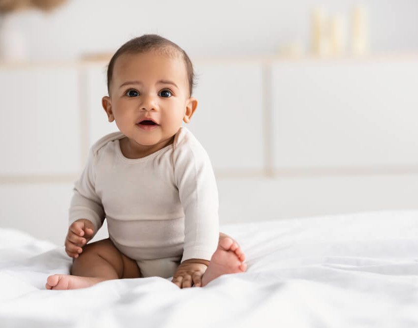 Foto que ilustra matéria sobre quarto de bebê simples mostra um bebê de poucos meses sentado em cima de uma cama olhando para a câmera. Ao fundo, desfocados, aparecem alguns móveis brancos.