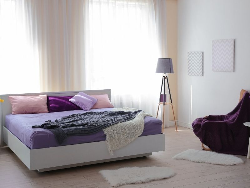 Quarto com cama e paredes em tonalidades lilás. Há também na imagem almofadas, travesseiros, uma poltrona, um lustre de chão, janelas amplas com cortina e dois tapetes pequenos brancos. 
