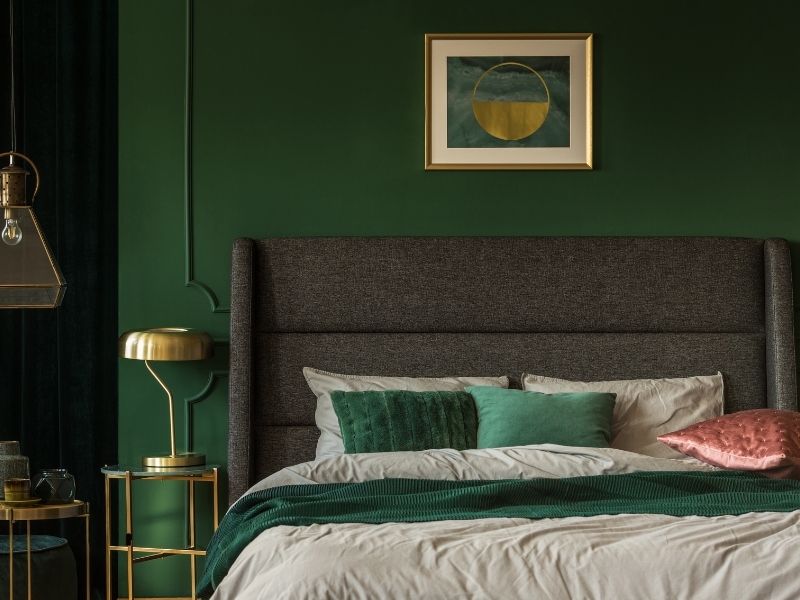 Quarto com parede verde, cama de casal com almofadas da mesma cor da parede. Há também na imagem abajur, lâmpadas e um quadro em tons dourados.