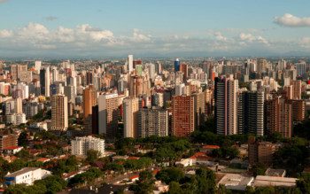 Matéria que fala sobre bairros de Curitiba mostra uma foto panorâmica da cidade pelo alto. Em primeiro plano, mais abaixo, há uma área verde, com muitas árvores. Um pouco mais atrás, diversos prédios altos. E ao fundo o céu azul com algumas poucas nuvens.