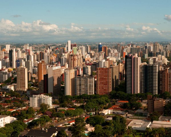 Matéria que fala sobre bairros de Curitiba mostra uma foto panorâmica da cidade pelo alto. Em primeiro plano, mais abaixo, há uma área verde, com muitas árvores. Um pouco mais atrás, diversos prédios altos. E ao fundo o céu azul com algumas poucas nuvens.