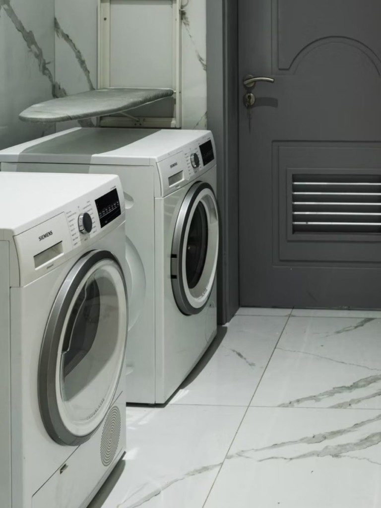A imagem mostra uma área de serviço pequena em tons de cinza. Nela há duas máquinas de lavar e uma tábua de passar roupa.