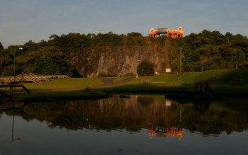 Foto que ilustra matéria sobre parques em Curitiba mostra uma panorâmica do Parque Tanguá, com um lago em primeiro plano e ao fundo uma grande pedreira onde se encontra, no topo, o mirante do parque