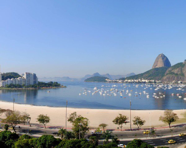 Foto que ilustra matéria sobre a Zona Sul do RJ mostra a Enseada de Botafogo, com carros passando na pista logo abaixo da imagem, uma faixa de areia, o espelho d’água com vários barcos e o Pão de Açúcar ao fundo.