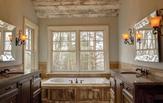 Banheiro rústico amplo com janelas largas, banheira revestida de madeira e pia de material em tom terroso