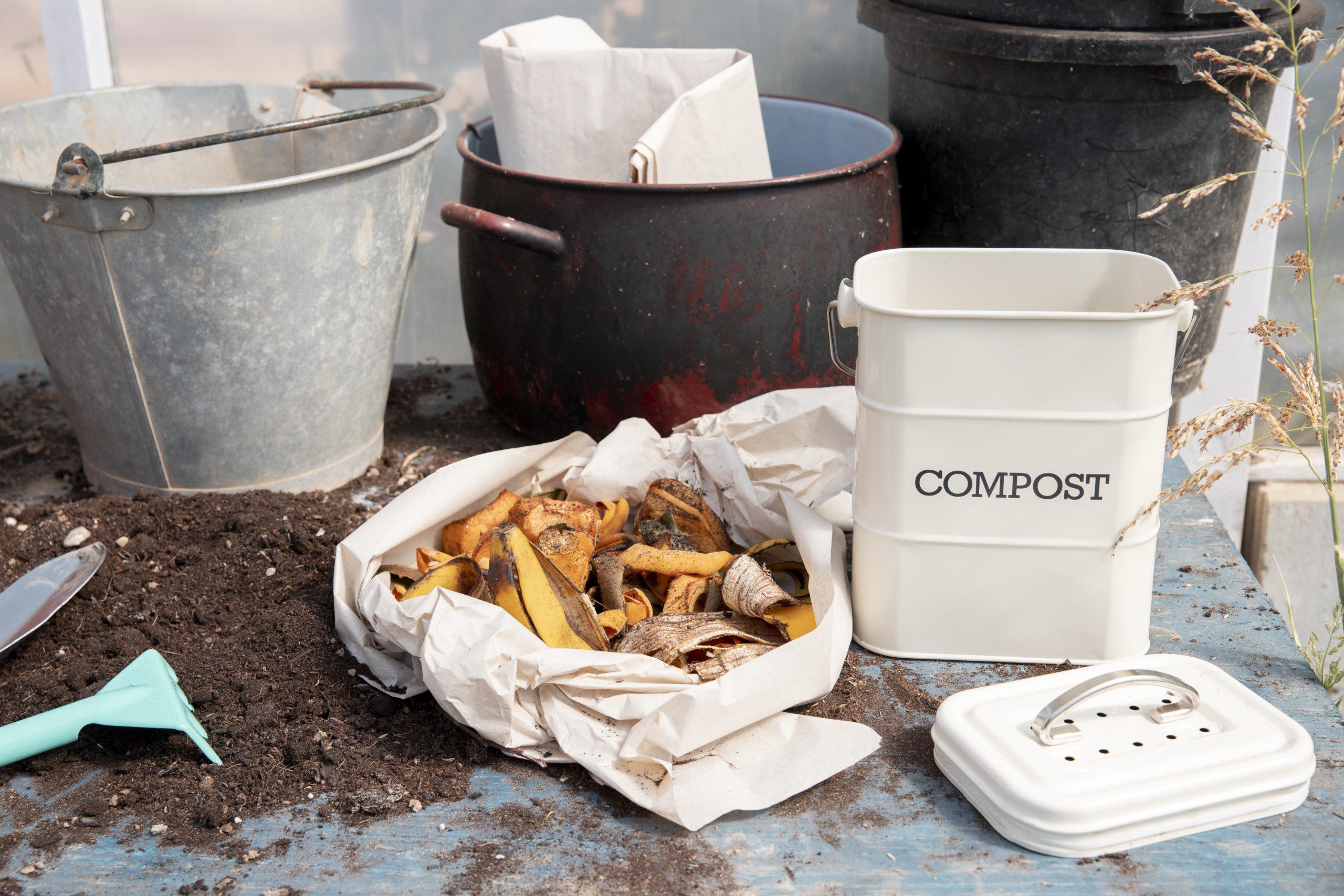 Imagem de um ambiente que está sendo preparado para a criação de uma composteira doméstica. Há uma mesa com baldes de aço no fundo, uma sacola com orgânicos e um balde de plástico que servirá de composteira. 