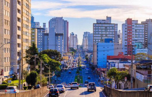Foto que ilustra matéria sobre custo de vida em Curitiba mostra uma grande avenida do centro da cidade, com carros passando na via ao centro e altos prédios em volta.