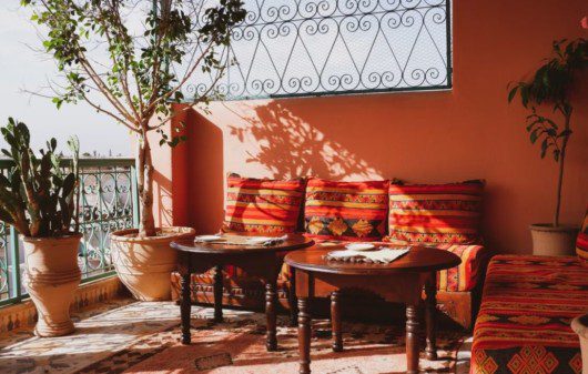 decoração de varanda com tons laranjas, sofás com almofadas listradas, mesas de centro, e a iluminação batendo em parte do ambiente. Há plantas decorando as beiras.