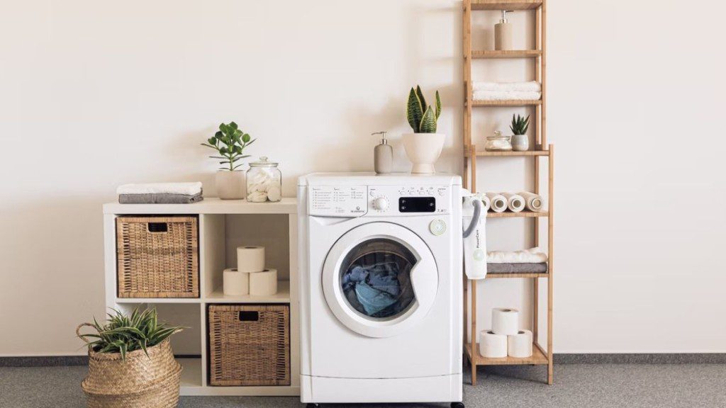 A imagem mostra uma área de serviço simples. Nela há uma máquina de lavar, um armário e uma estante em cada um dos lados dela. Há na imagem itens como toalhas, papel higiênico e plantas.