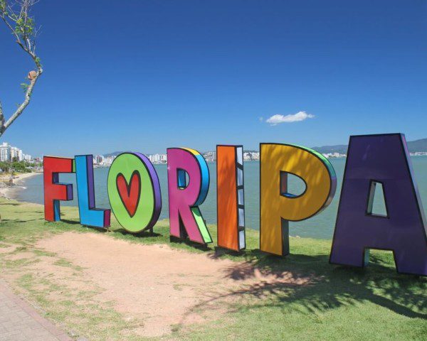 Foto que ilustra matéria sobre o que fazer em Florianópolis mostra em detalhes um letreiro com a palavra “Floripa” escrita em letras coloridas à beira de uma das praias da cidade.