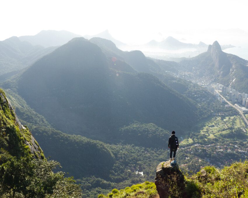 Imagem panorâmica de um homem de costas em cima da Pedra da Gávea olhando as montanhas do Rio de Janeiro para ilustrar matéria sobre parques no Rio de Janeiro