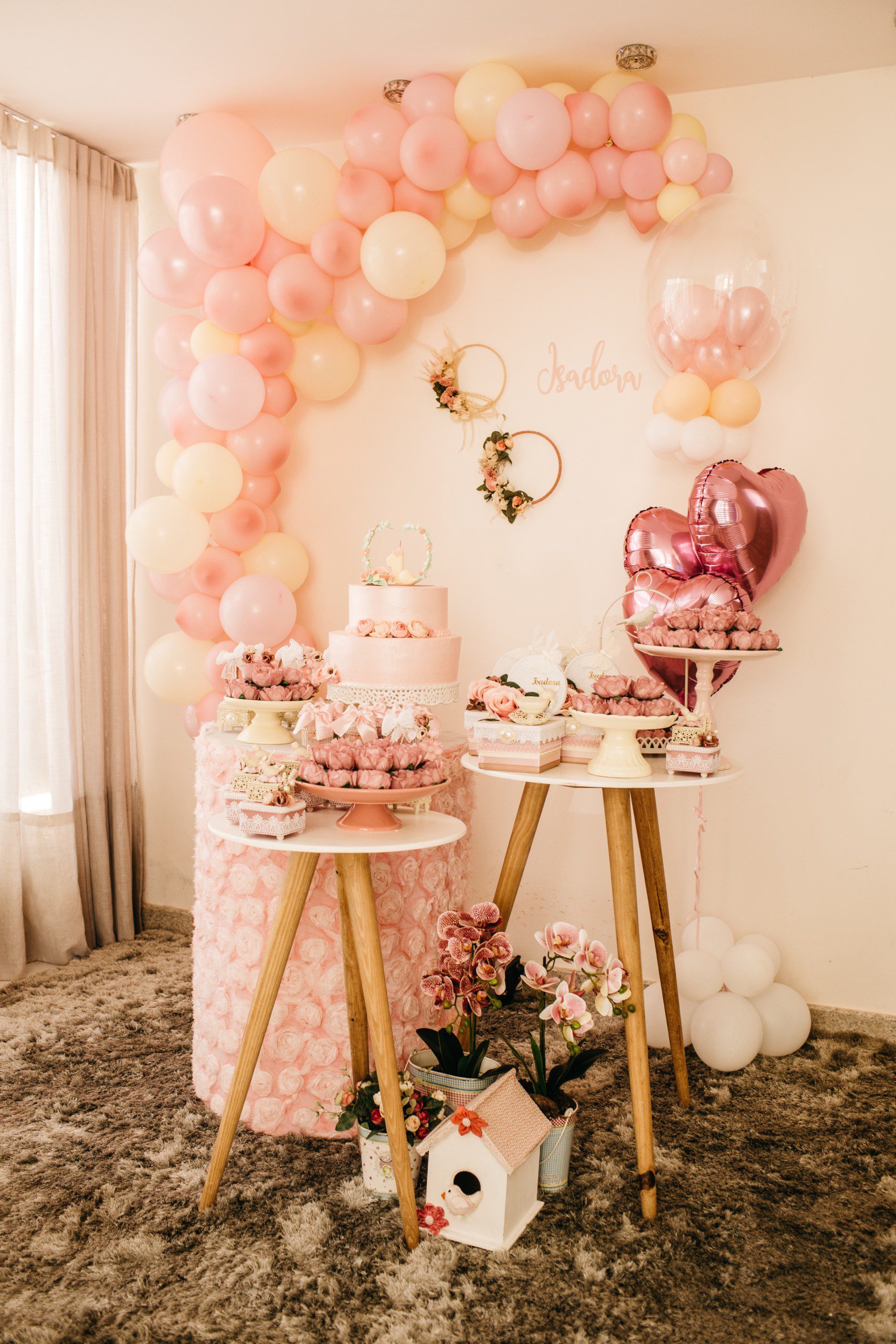 Imagem de uma decoração de festa simples em tons de rosa com arco de balões desconstruído, quadrinhos na parede, arranjos de flores e bolo de andares. 