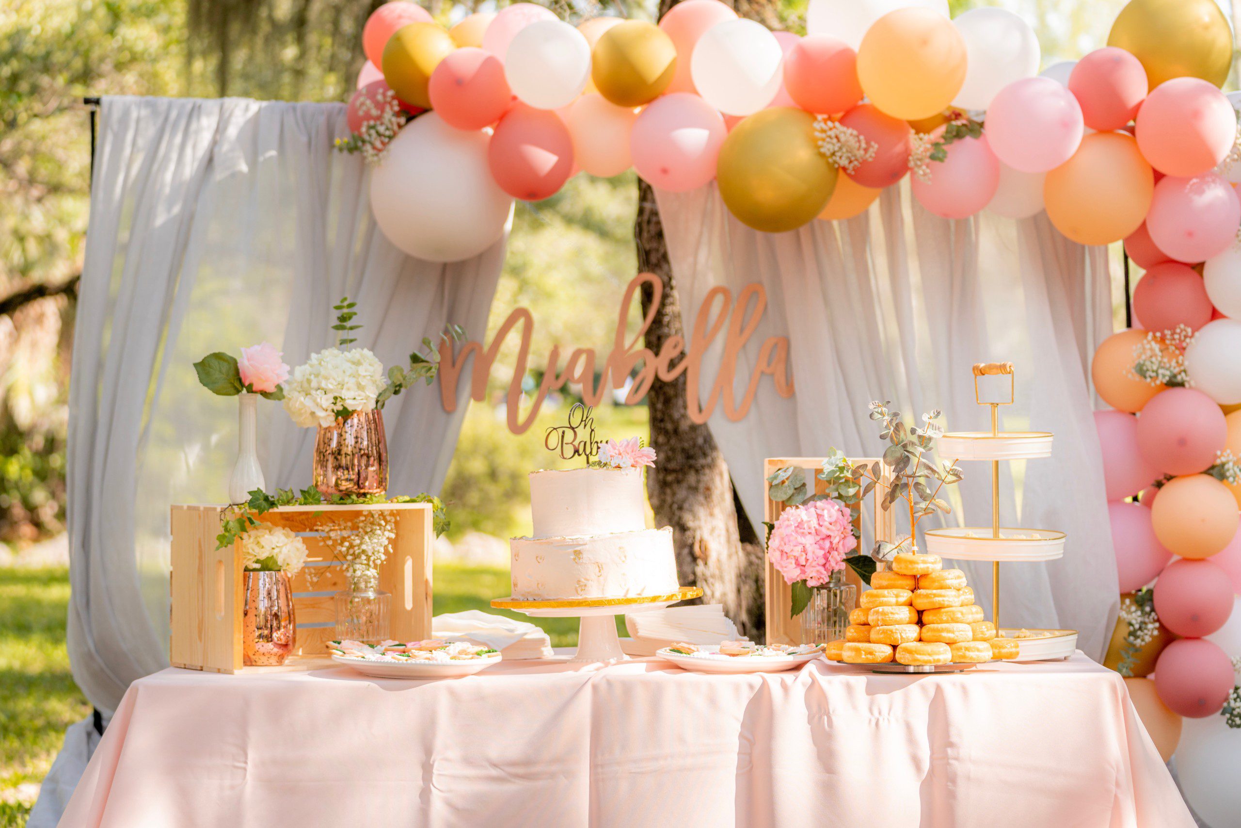 Imagem de uma decoração de festa simples em tons pastéis com um arco de balões desconstruído, mesa de doces com bolo de dois andares e vasos de flores.