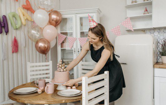 Imagem de uma moça arrumando o bolo com uma decoração de festa simples com balões em tons de rosa e rose gold.
