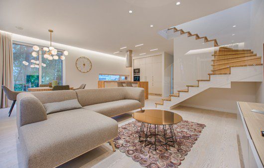 Foto que ilustra matéria sobre ideias de itens para casa mostra uma grande sala de estar com um sofá em cor neutra, mesa de centro, tapete e escada de madeira ao fundo