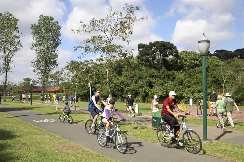 Foto que ilustra matéria sobre ciclovias em Curitiba mostra uma pista de asfalto repleta de ciclistas em meio a um parque arborizado.