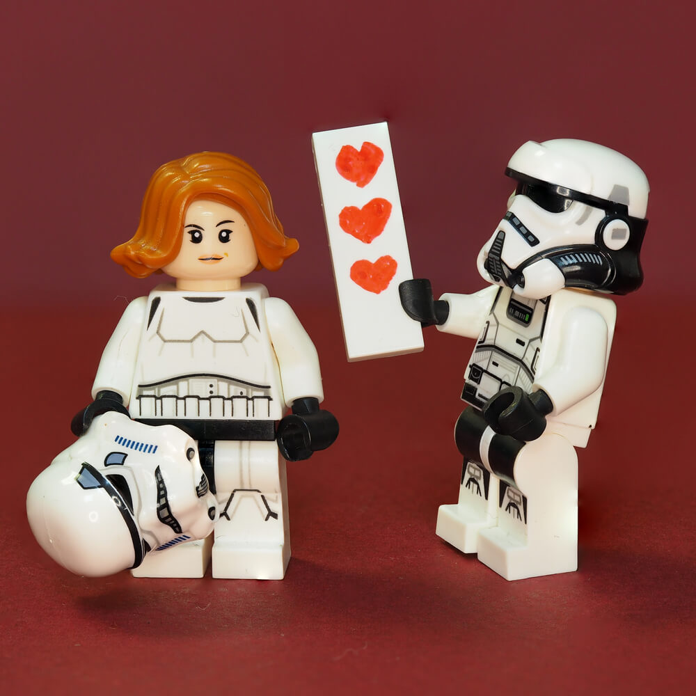 Dois bonecos de lego do filme Star Wars. O masculina dá um cartão com corações para o feminino. Eles simulam um casal.