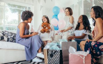 Foto que ilustra matéria sobre decoração para chá de bebê mostra cinco mulheres sentadas em dois sofás. Elas estão sorridentes e cercadas de presentes. A segunda mulher da esquerda para direita está vestida de branco e grávida e segura um ursinho de pelúcia. No centro da imagem, alguns balões nas cores azul, branco e rosa decoram o ambiente.
