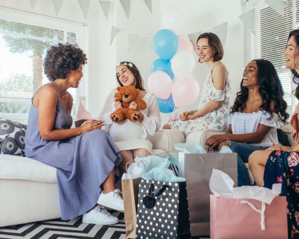 Foto que ilustra matéria sobre decoração para chá de bebê mostra cinco mulheres sentadas em dois sofás. Elas estão sorridentes e cercadas de presentes. A segunda mulher da esquerda para direita está vestida de branco e grávida e segura um ursinho de pelúcia. No centro da imagem, alguns balões nas cores azul, branco e rosa decoram o ambiente.