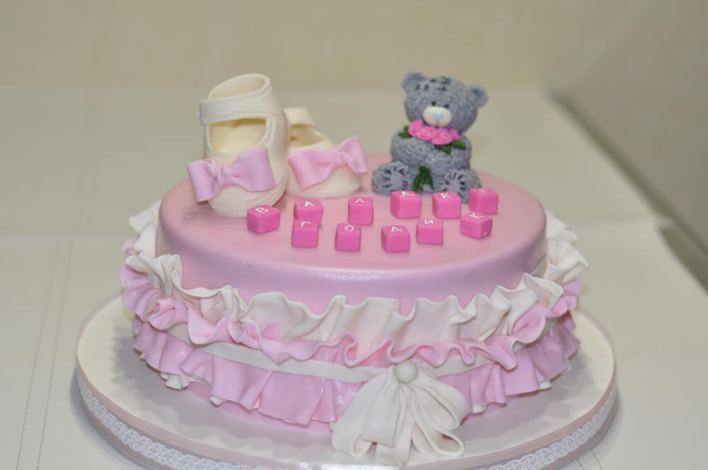 A imagem mostra um bolo redondo, em tons de rosa e branco, com um ursinho em cima. Há também letras e um sapatinho no bolo.