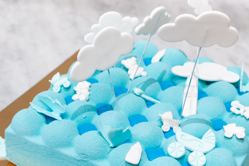 A imagem mostra um bolo em formato retangular com uma cobertura de nuvens. Em cima do bolo há desenhos como ursinhos,mamadeira, carrinho de bebê e nuvens.